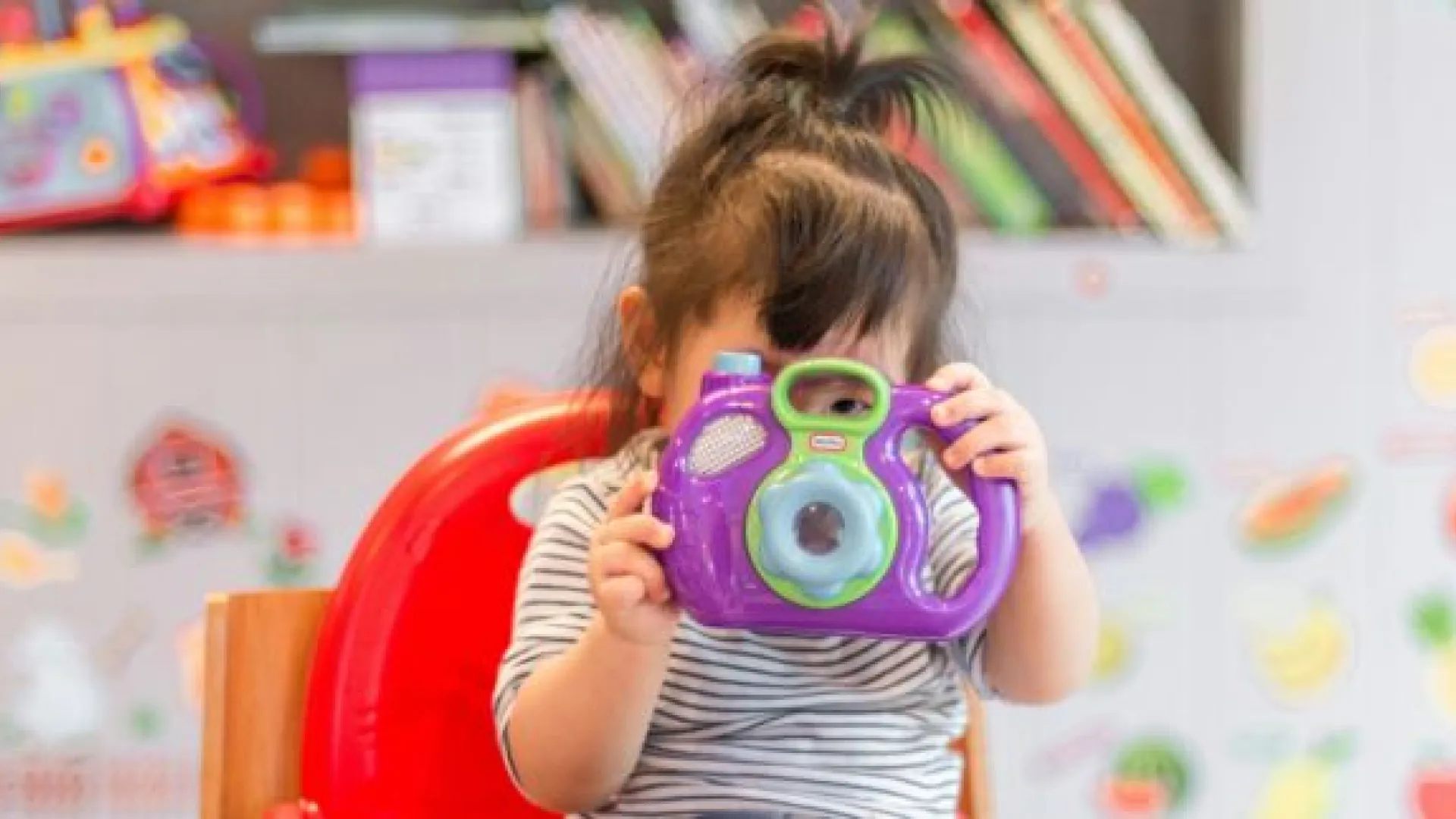 little girl holding up a toy camera in a classroom