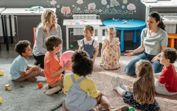 Two teachers instructing elementary students seated in a circle on the floor