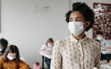 educator wearing mask and holding books standing in front of students at desks