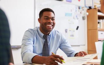 male teacher seated at desk smiling and pointing to documents