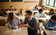 Male teacher kneeling down to work with young student at her desk