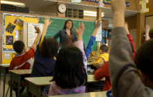 teacher standing at front of middle school classroom with students raising their hands