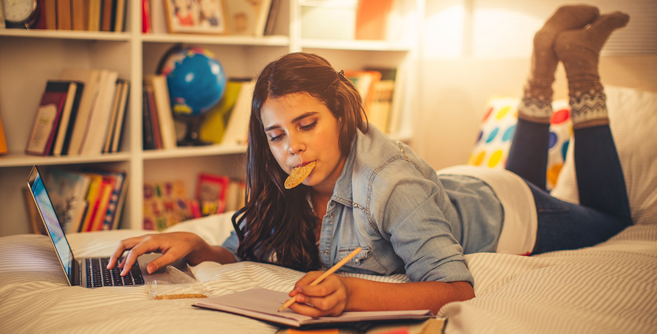 Image of teenager lying on bed eating a cookie and doing homework